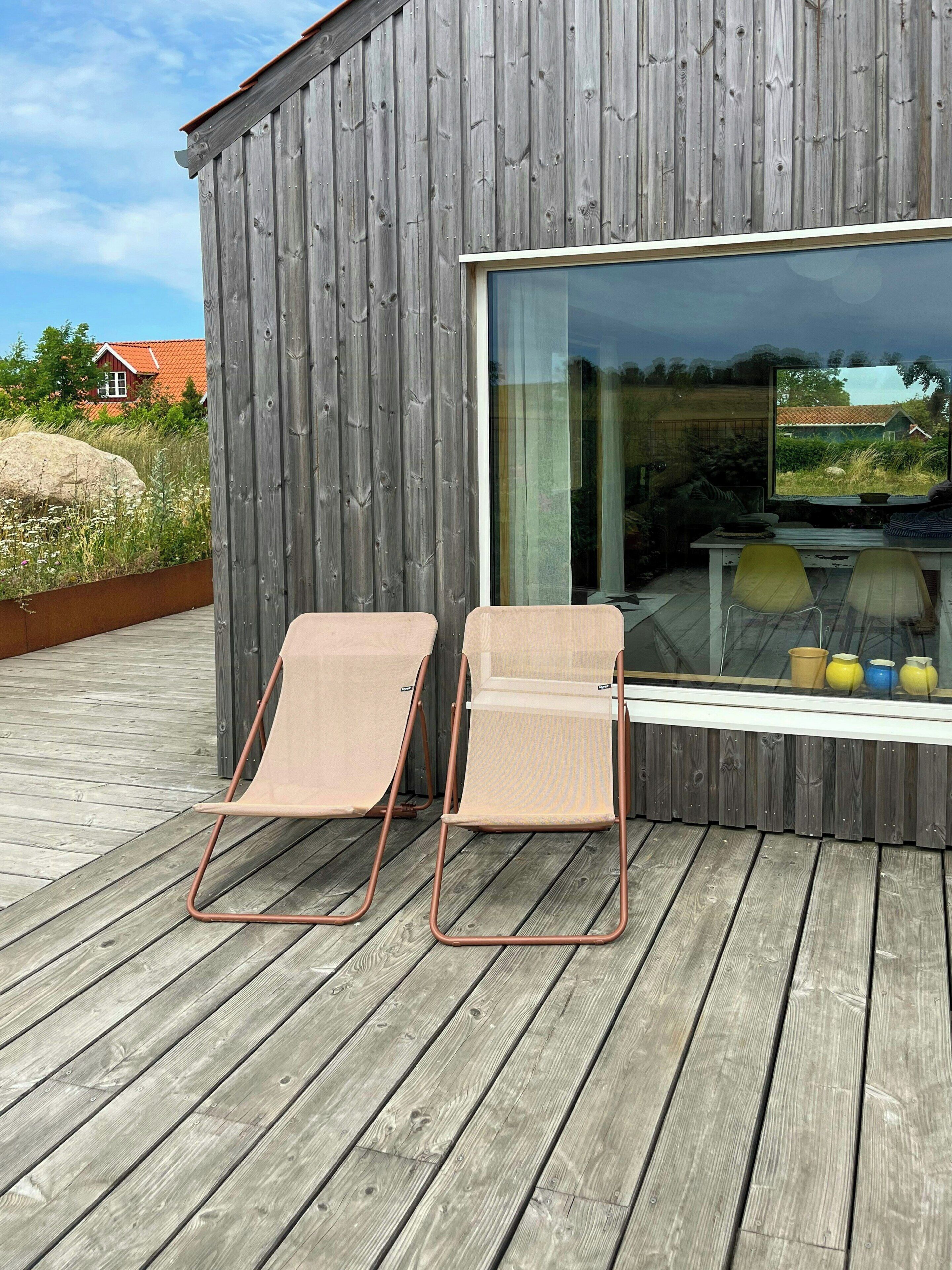 Une terrasse en bois avec deux fauteuils de jardin beiges et une porte vitrée donnant sur une pièce.