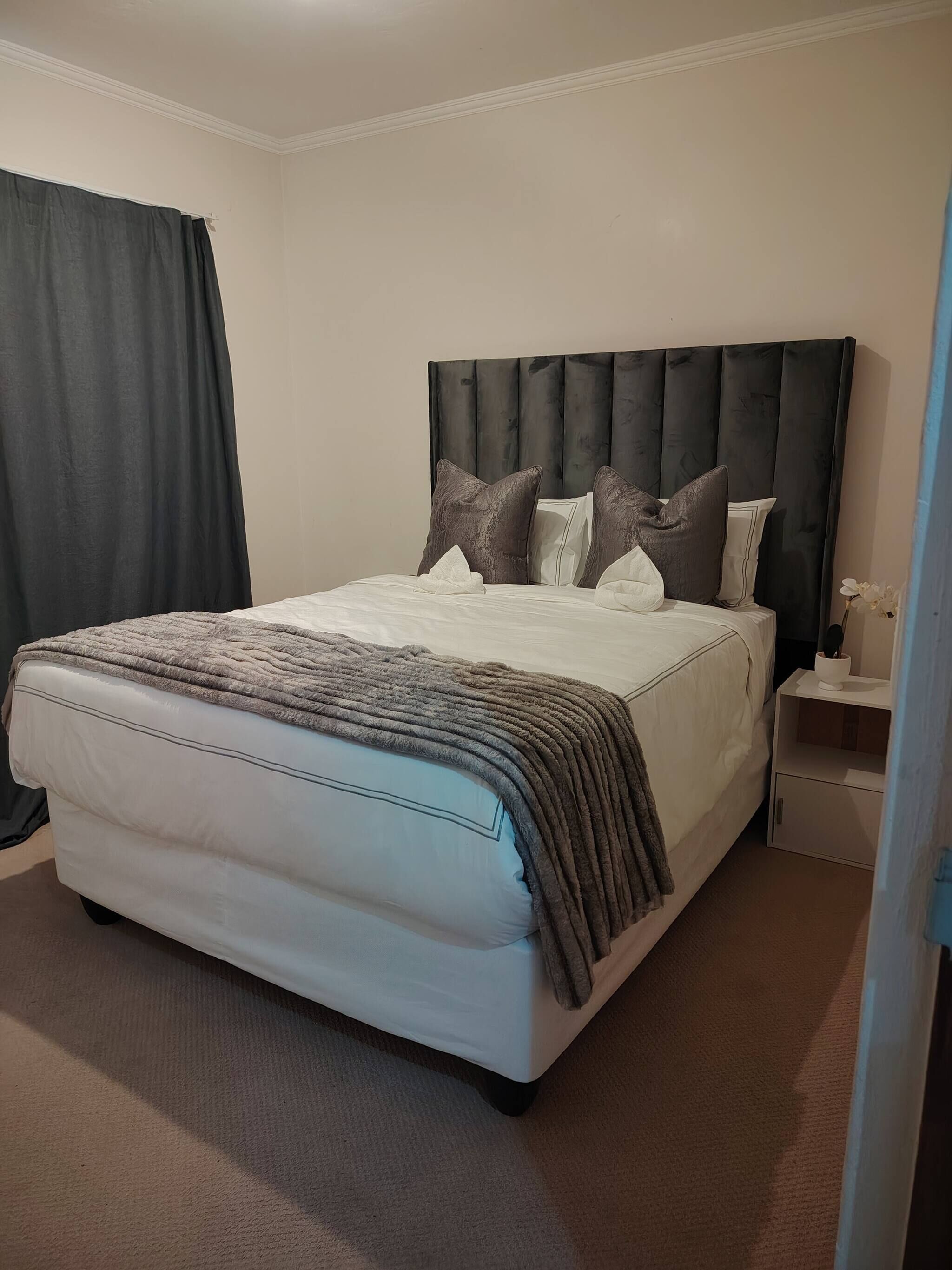 A neatly made bed with a grey and white duvet, a dark headboard, and a bedside table with a vase of flowers.