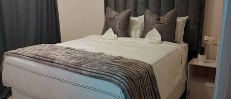 A neatly made bed with a grey and white duvet, a dark headboard, and a bedside table with a vase of flowers.