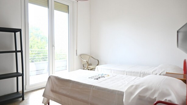 A hotel room with two beds, a chair, a shelf, and a sliding glass door to the outside. - SE293 - Senigallia, two-room apartment just steps from the sea and the center of Senigallia. (Senigallia)