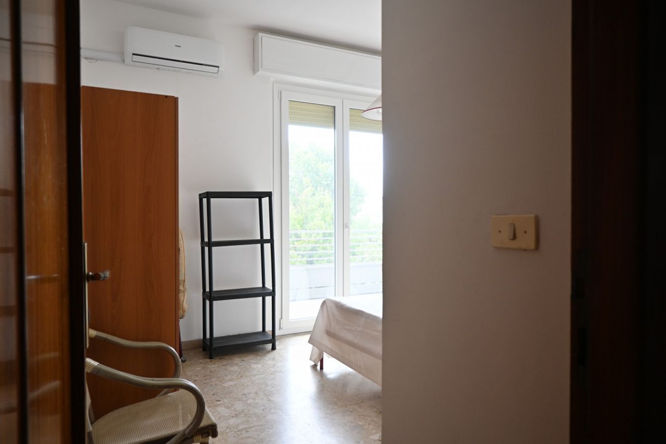 A bedroom with a bed, a wooden wardrobe, a black shelf unit, and a window with a view of greenery.