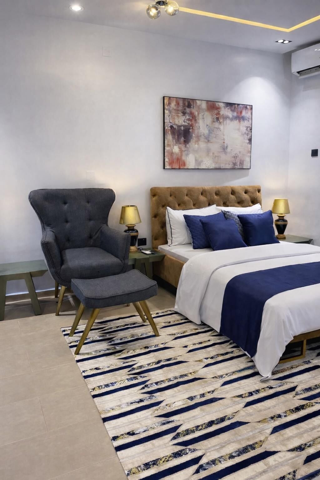 A bedroom with a tufted headboard bed, a grey armchair, a patterned rug, and a modern abstract painting.