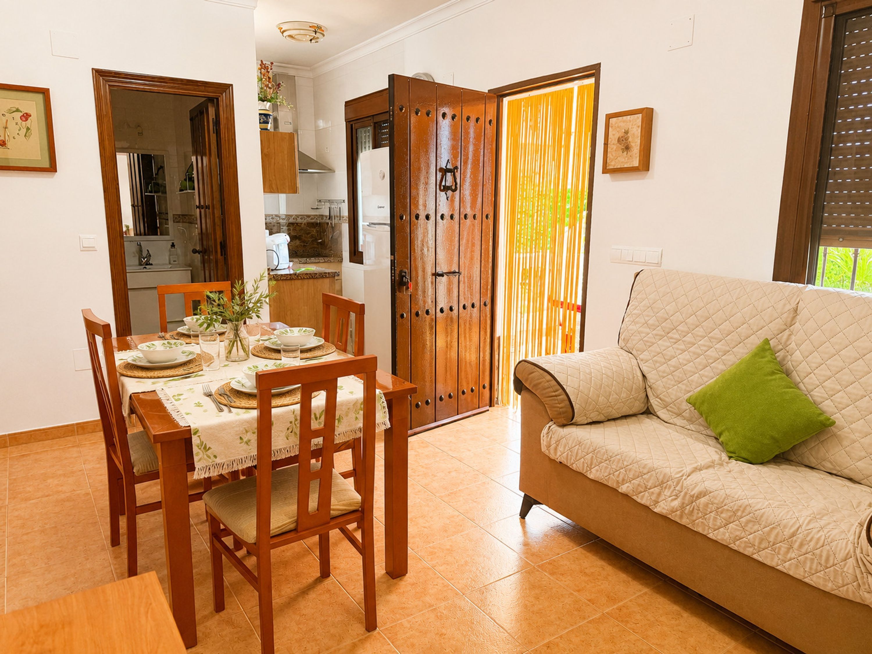 Un comedor con una mesa preparada para cuatro personas, un sofá con un cojín verde y una puerta corredera que da a la cocina.