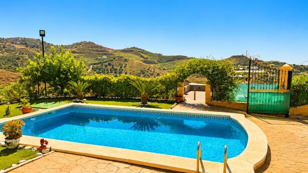 A swimming pool with a hexagonal tile border, surrounded by a garden with various plants and a view of hills in the background. - Cortijo las Flores con Piscina Privada (Torrox)
