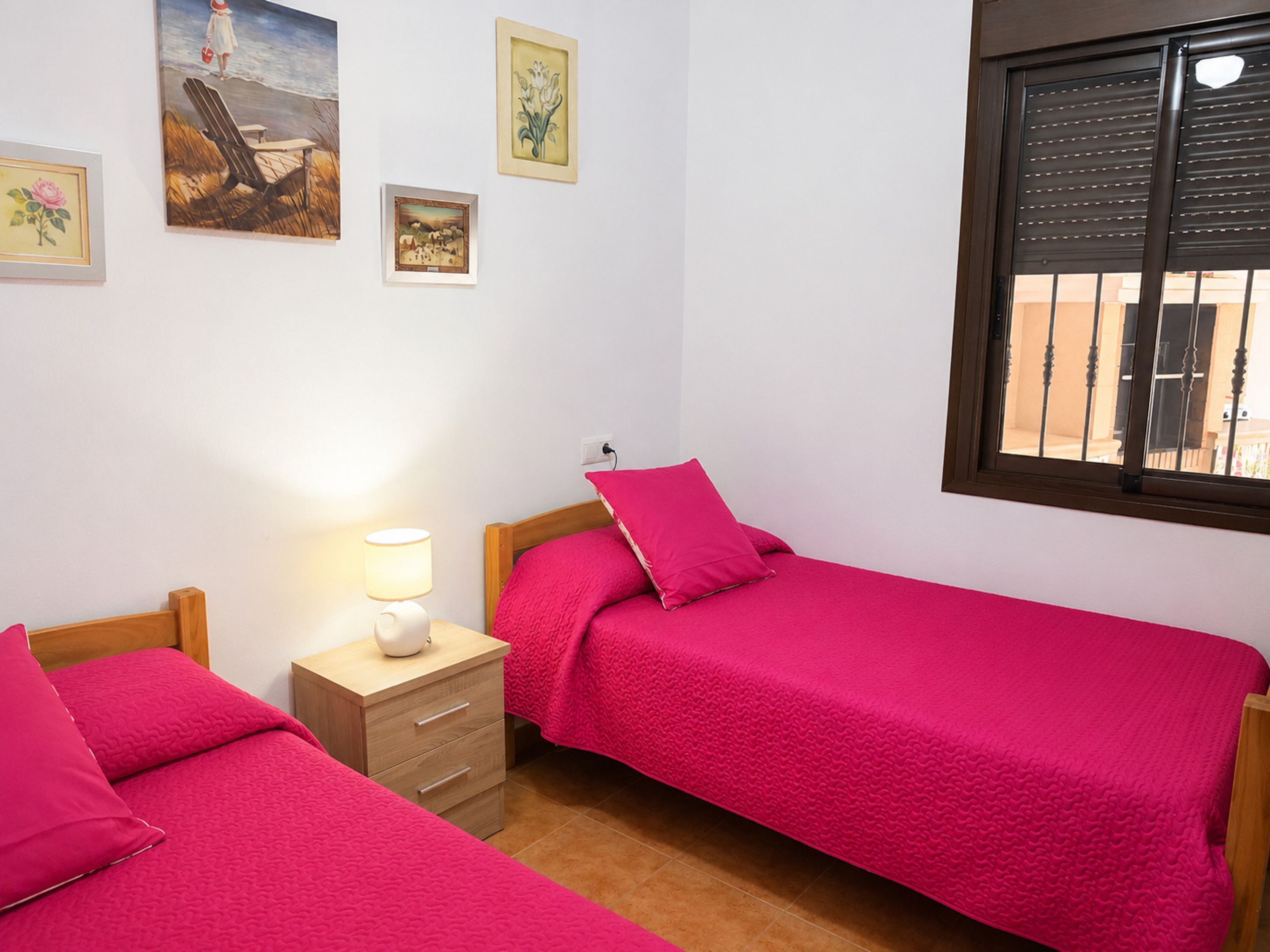 A bedroom with two beds, a bedside table with a lamp, and a window with blinds.