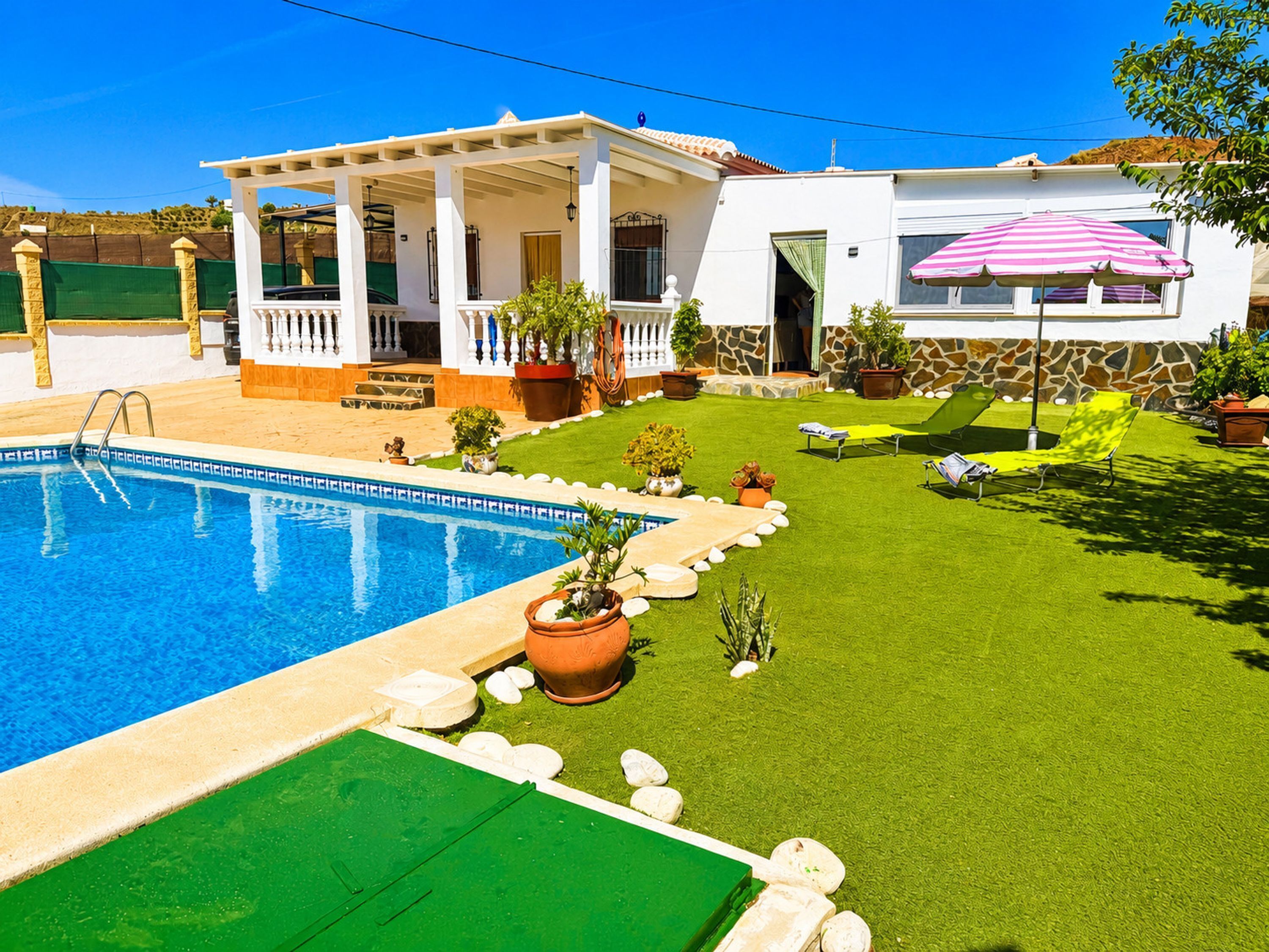 Una piscina, un porche cubierto y un jardín con mobiliario exterior.