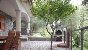 Un patio exterior con suelo de piedra, sillas de madera, una mesa, un muro de piedra y una parrilla de ladrillo.