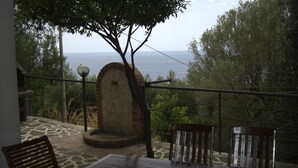 Una terraza con una mesa y sillas de piedra, un banco de piedra, un árbol y vista al mar.