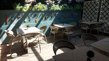 Uma área externa com mesas e cadeiras, cercada por plantas decorativas e obras de arte na parede.
