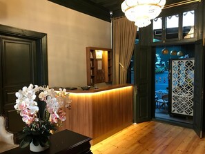Un mostrador de recepción con detalles de madera, una araña de cristal y vistas al exterior a través de una puerta con celosías decorativas.