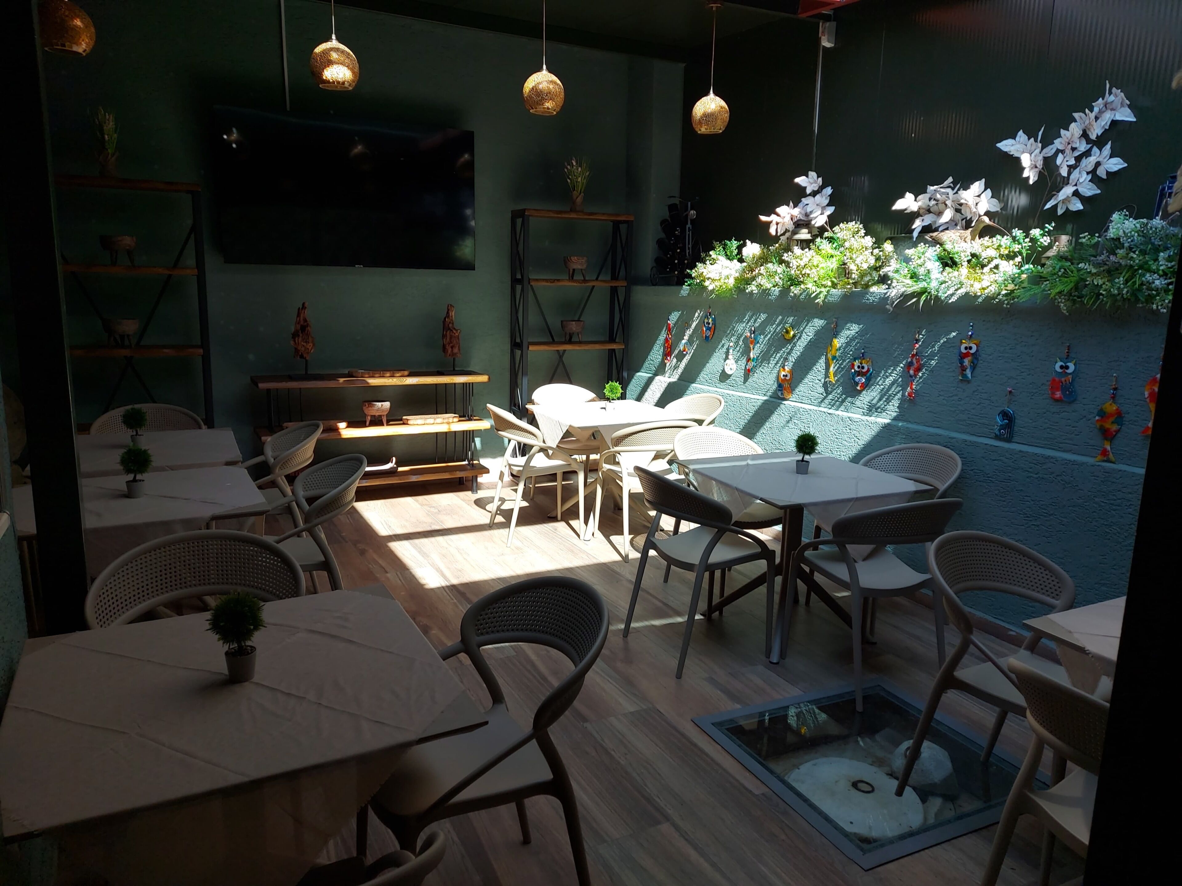 Interior de restaurante com várias mesas e cadeiras, uma TV de tela plana, luminárias pendentes e uma parede decorada com diversos enfeites.