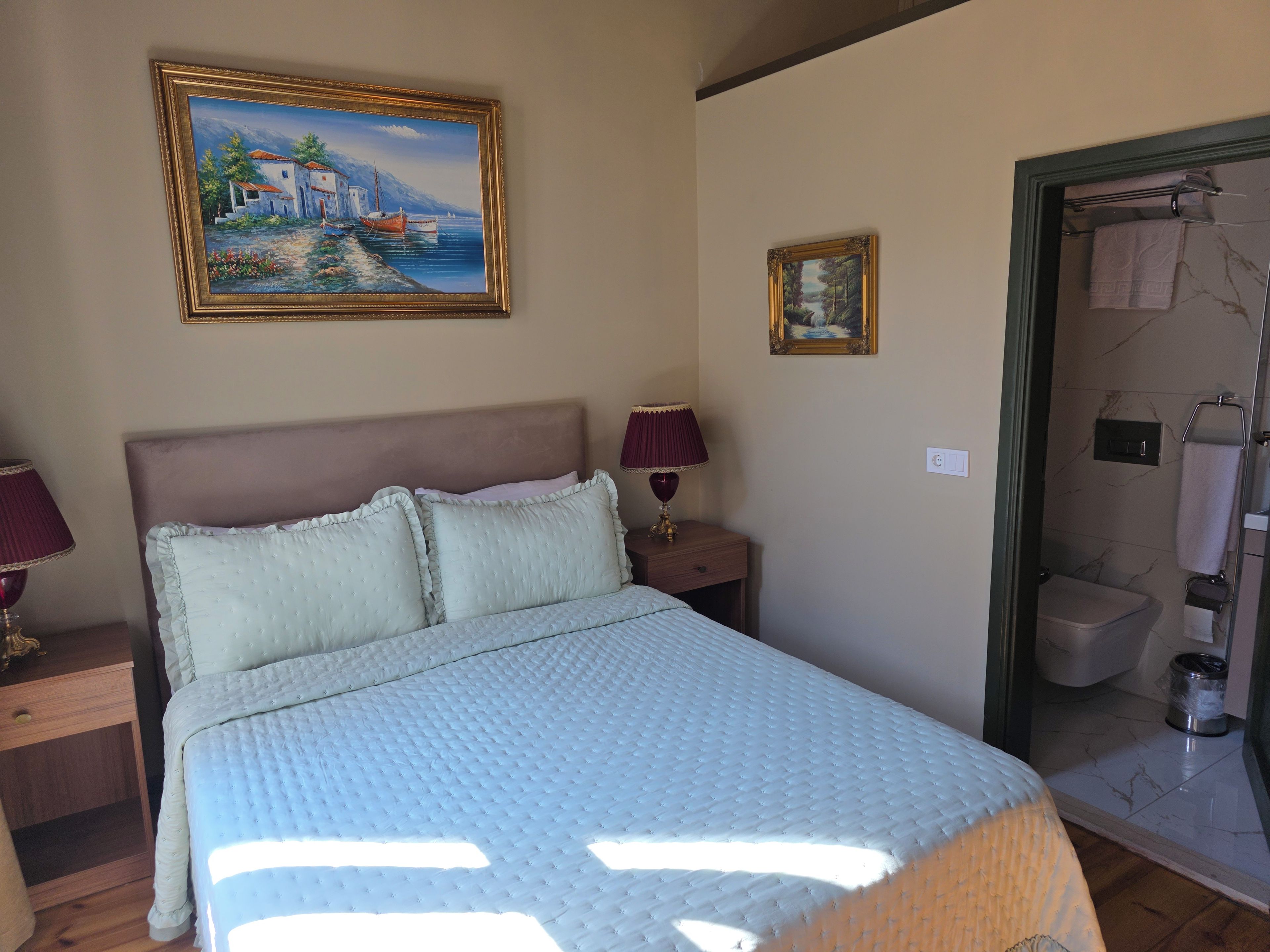 Una cama bien tendida con almohadas, una mesita de noche con lámpara, un cuadro en la pared y la puerta del baño.