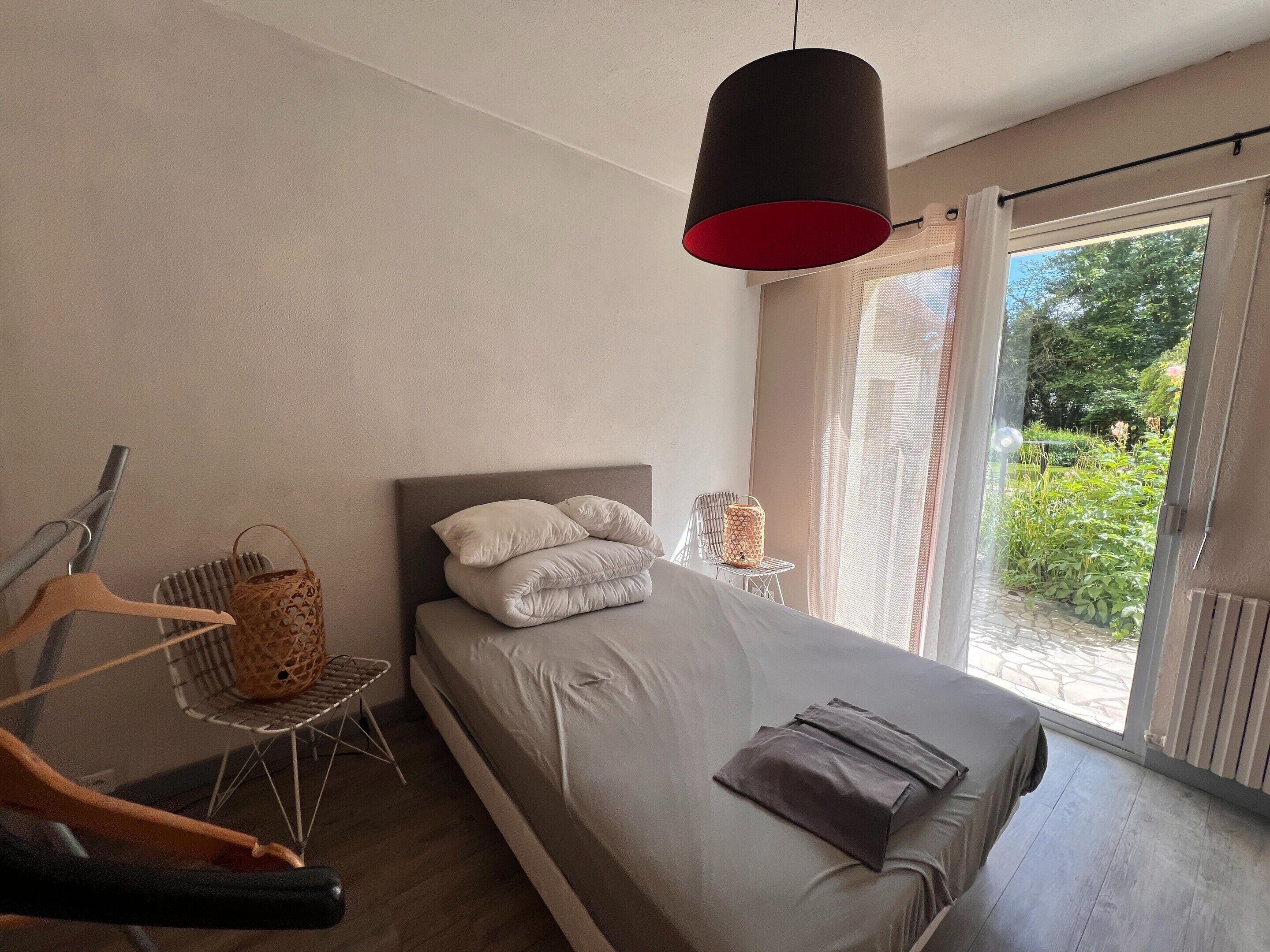 A bedroom with a bed, a hanging light fixture, a chair, and a window with a view of greenery.