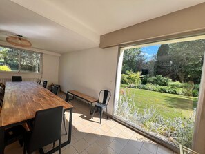 A spacious room with a large dining table, chairs, a bench, and a view of a well-maintained garden through large windows. - Grand French Family Holiday Home (Saint-Germain-sur-Bresle)