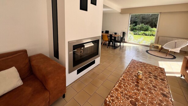 A modern living room with a fireplace, a dining area, and a view of a garden. - Grand French Family Holiday Home (Saint-Germain-sur-Bresle)