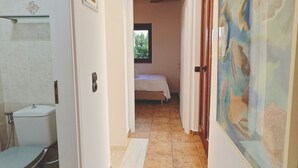 A hallway with a bathroom on the left, a bed in the center, and a painting on the right. - Emmelia Stone Maisonette: Panoramic Sea View (Kalamata)