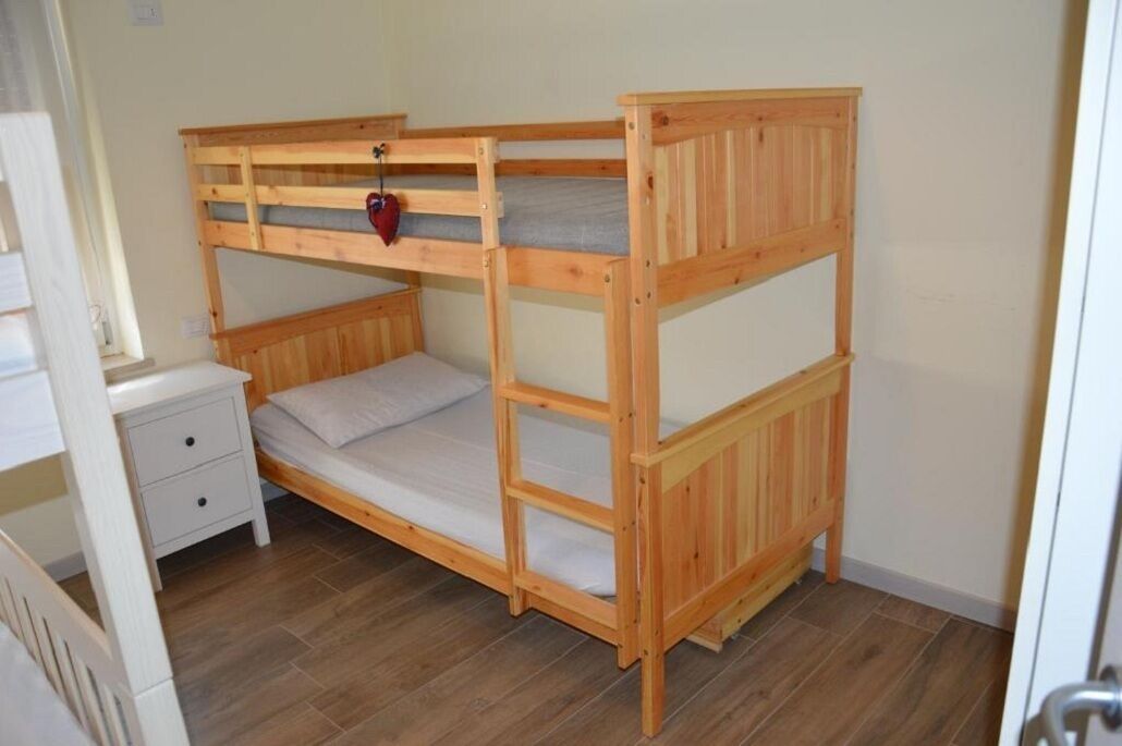Un letto a castello con un comodino bianco e cassetti, pavimento in legno e una parete semplice.