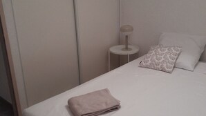 Una cama bien hecha con una toalla doblada, una lámpara de noche y una almohada estampada sobre un colchón blanco en una habitación con una puerta cerrada y una ventana.