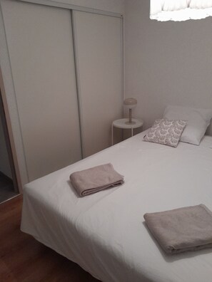 A neatly made bed with a folded towel, a bedside lamp, and a patterned pillow on a white mattress in a room with a closed door and a window. - A cozy home with a yard near downtown, in a quiet neighborhood. 5 minutes from the lake, 8 minutes from the tram (Le Crès)
