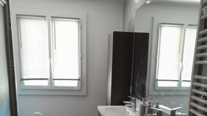 Un baño con un lavamanos blanco, tocador oscuro y un espejo grande. Hay dos ventanas con persianas, un bote de basura blanco y una alfombra negra en el suelo.