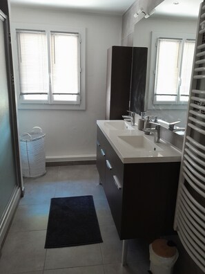 A bathroom with a white sink, dark vanity, and a large mirror. There are two windows with blinds, a white trash bin, and a black rug on the floor. - A cozy home with a yard near downtown, in a quiet neighborhood. 5 minutes from the lake, 8 minutes from the tram (Le Crès)