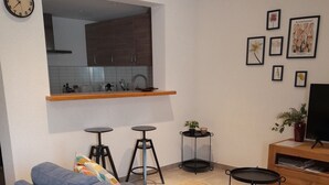 Una sala de estar moderna con un sofá azul, una pequeña cocina y un televisor de pantalla plana sobre un soporte de madera.