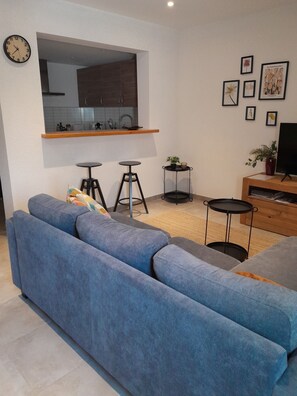 A modern living room with a blue sofa, a small kitchen area, and a flat-screen TV on a wooden stand. - A cozy home with a yard near downtown, in a quiet neighborhood. 5 minutes from the lake, 8 minutes from the tram (Le Crès)