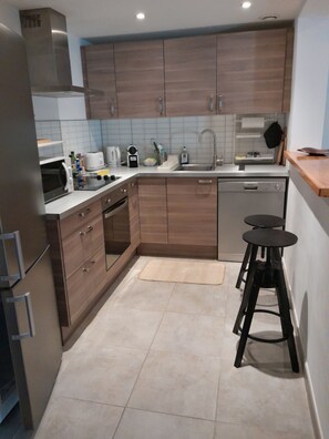 A modern kitchen with wooden cabinets, a stainless steel range hood, a dishwasher, a microwave, and a coffee maker. - A cozy home with a yard near downtown, in a quiet neighborhood. 5 minutes from the lake, 8 minutes from the tram (Le Crès)