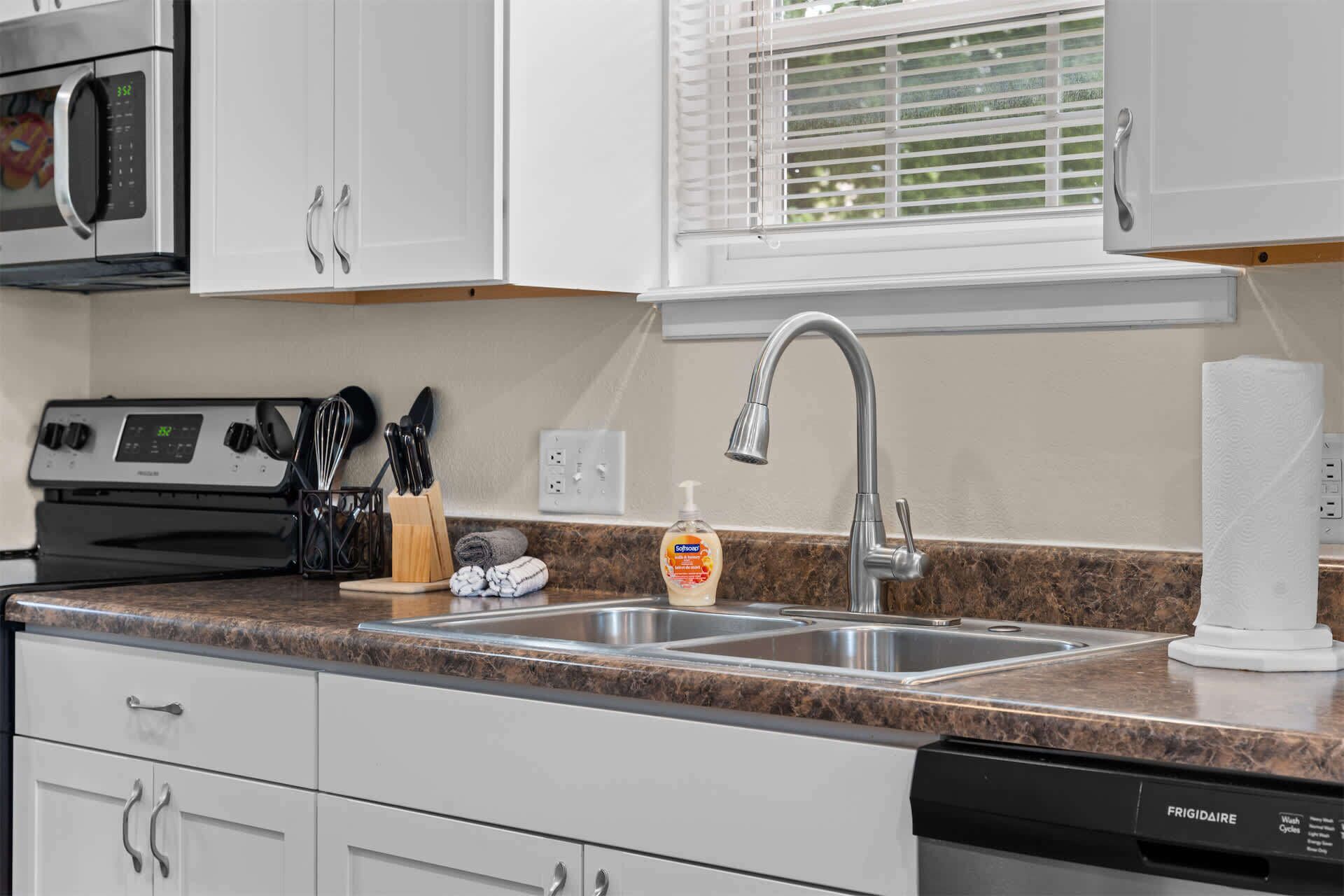 Una cocina con armarios blancos, un lavamanos de acero inoxidable, un microondas, una estufa y una botella de detergente.