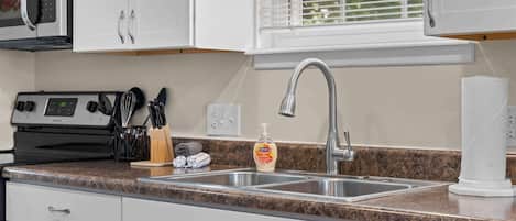 Una cocina con armarios blancos, un lavamanos de acero inoxidable, un microondas, una estufa y una botella de detergente.