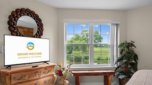 A room with a television displaying a welcome message, a large window with a view of greenery, a wooden bench, and a dresser with drawers. - Pond View Escape - Resort Access (Branson)