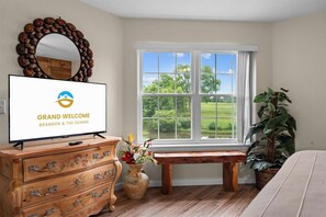 Ein Zimmer mit Fernseher, der eine Willkommensbotschaft zeigt, einem großen Fenster mit Blick ins Grüne, einer Holzbank und einer Kommode mit Schubladen.