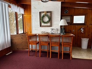 A wooden interior with a table and chairs, a welcome sign, a wall-mounted wreath, and a window with blinds.