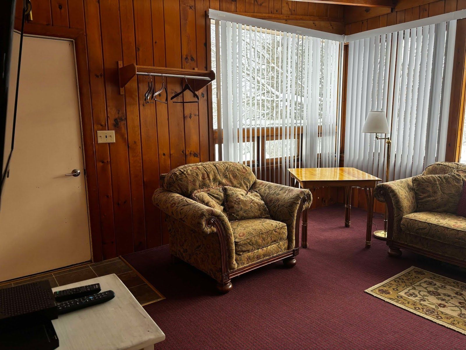 A room with wooden walls, a patterned armchair, a sofa, a small table, and a lamp.