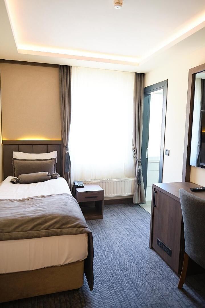 Een hotelkamer met een bed, nachtkastje, bureau, stoel en een groot raam met gordijnen.
