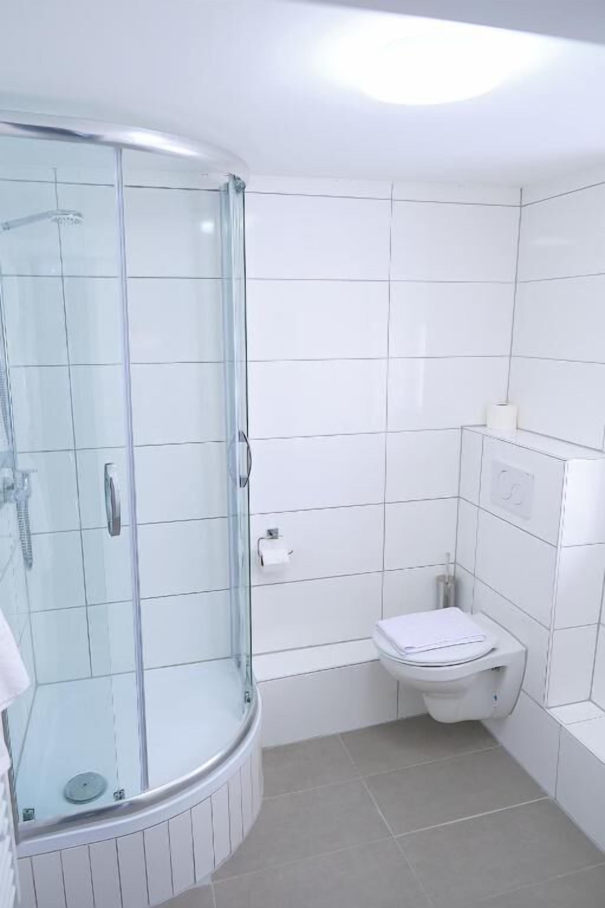 Et badeværelse med en glasafskærmet bruseniche, toilet og hvide flisevægge.