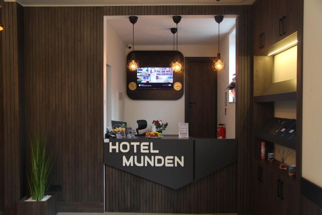 Ein Hotelrezeptionstresen mit einem digitalen Bildschirm, zwei Pendelleuchten und einem Schild mit der Aufschrift 'HOTEL MUNDEN'.
