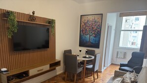 Ein Wohnzimmer mit Flachbildfernseher an einer Holzwand, einem Esstisch mit Stühlen, einer Couch und einem Baumgemälde.