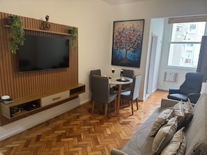 A living room with a flat-screen TV mounted on a wooden wall, a dining table with chairs, a sofa, and a painting of a tree. - Cozy Apartment - Copacabana/RJ 5min from the Beach (Rio de Janeiro)