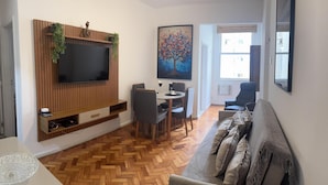 Ein Wohnzimmer mit Fernseher, Esstisch, Stühlen, einer Couch und einem Bild an der Wand.