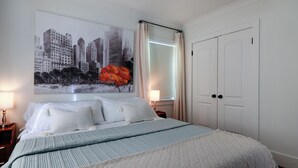 A bedroom with a bed, a nightstand, a wall-mounted lamp, a door, and a large cityscape painting. - Beautiful Avondale Bungalow, Downtown BHAM a 5 min drive, UAB & Airport 10 mins. (Birmingham)