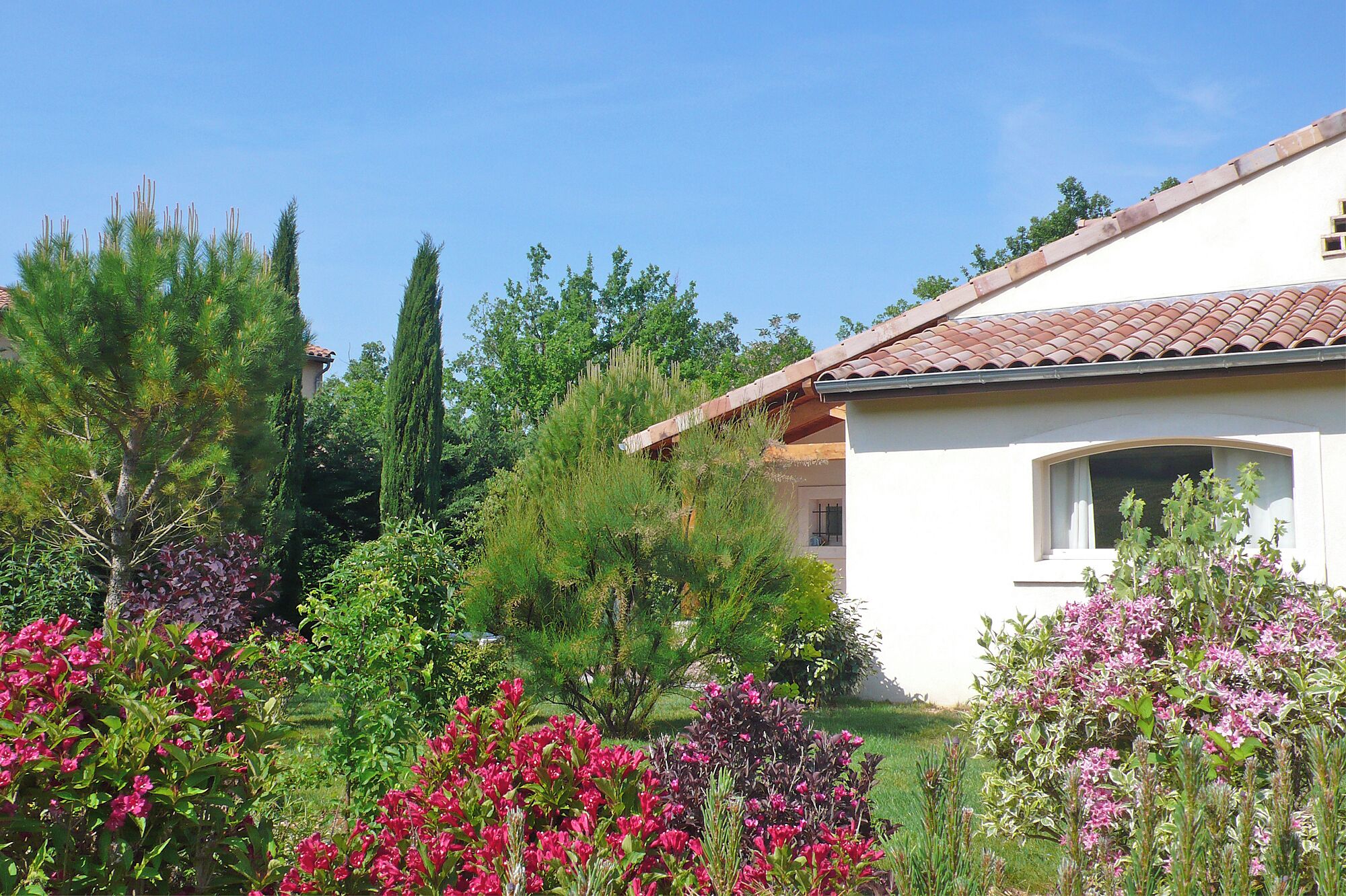 Un bâtiment blanc surmonté d’un toit recouvert de tuiles rouges, entouré d’une végétation luxuriante et de plantes en fleurs.