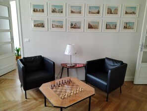 Ein Zimmer mit Schachbrett, zwei schwarzen Sesseln, einer Lampe und gerahmten Bildern an der Wand.