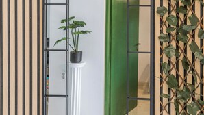 Et spejl med sort ramme viser et rum med en grøn dør, en hvid søjle og en potteplante.