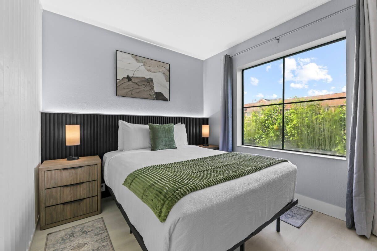A modern bedroom with a large bed, a nightstand, a wall-mounted lamp, a framed artwork, and a window with a view of greenery.
