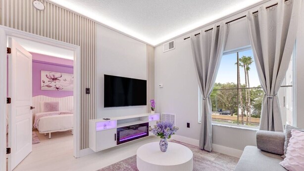A modern living room with a flat-screen TV, a white entertainment unit, a round coffee table with a vase, a sofa, and a view of palm trees through the window. - Disney Area Suite w/ Pool & Full Kitchen (Orlando)