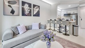 A modern living room with a grey sofa, a coffee table with a vase, and a kitchen area with bar stools. - Disney Area Suite w/ Pool & Full Kitchen (Orlando)