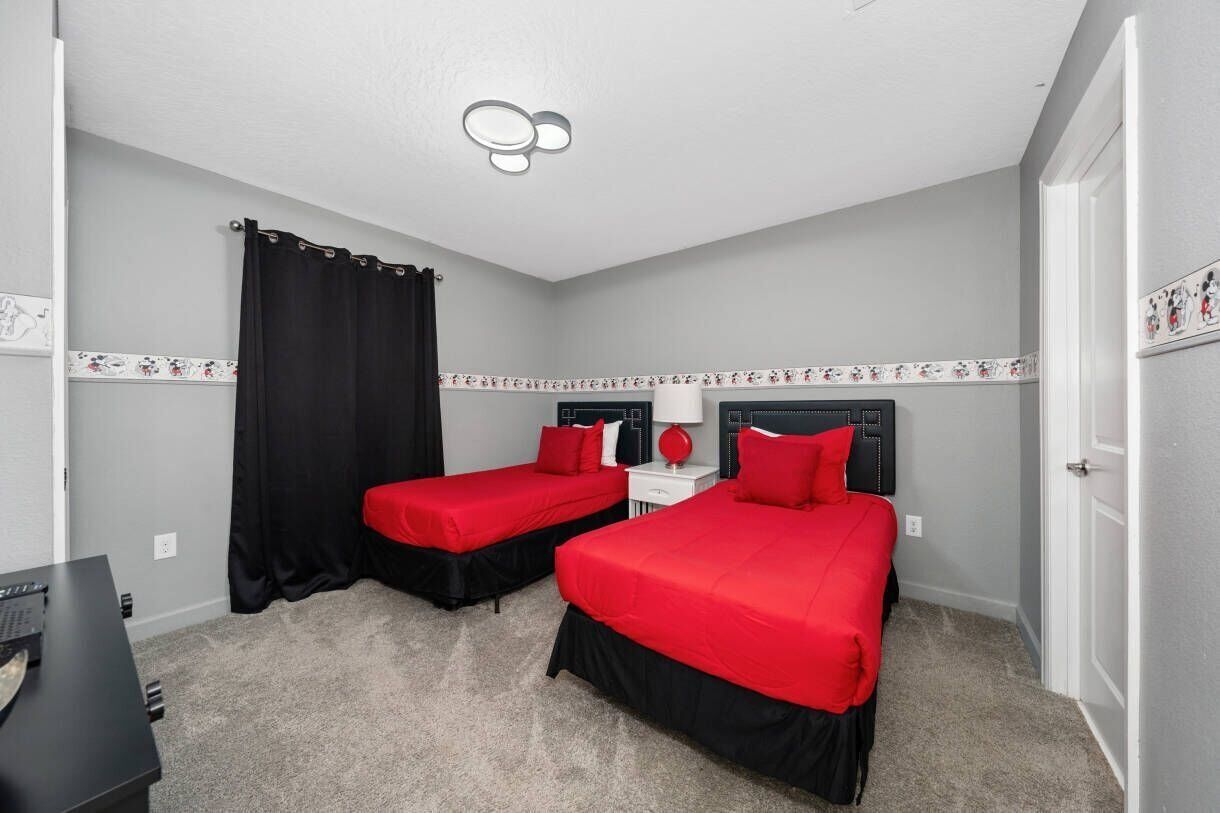 A bedroom with two beds, a desk, a chair, a ceiling light, and a door.