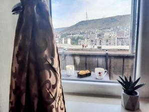 Une fenêtre donnant sur un paysage urbain, une plante en pot et un plateau avec des biscuits et une tasse.