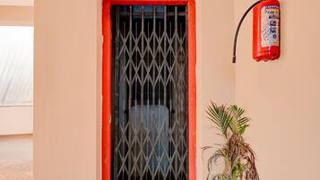 Une fenêtre avec une grille en métal et un cadre rouge, un extincteur fixé au mur, et une plante en pot placée à côté de la fenêtre.